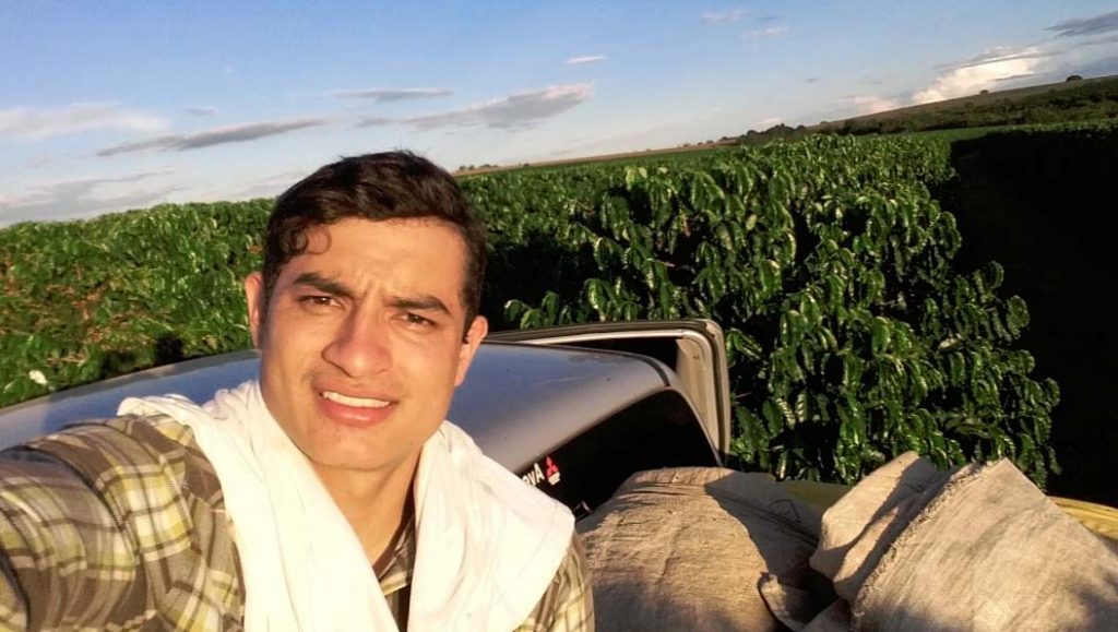 Heiber Andres Trujillo Samboni in the coffee fields. Selfie of a young man that is standing of the back of a pickuptruck full of bags. In the background you can see coffee plants.